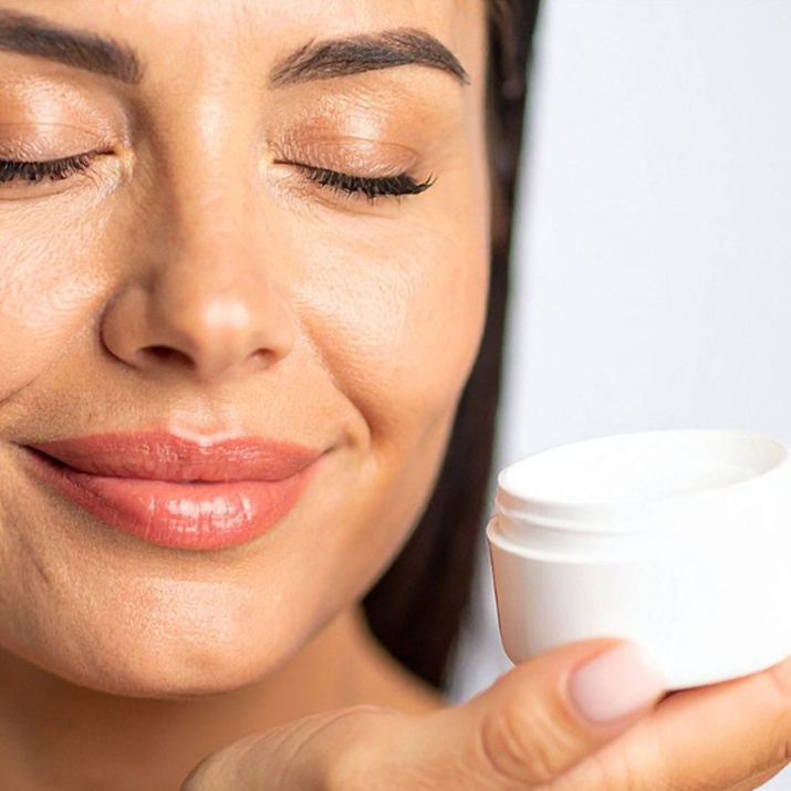 Primer plano de una mujer sonriente con los ojos cerrados sosteniendo un tarro de crema cosmética blanca, representando la experiencia sensorial y el bienestar.